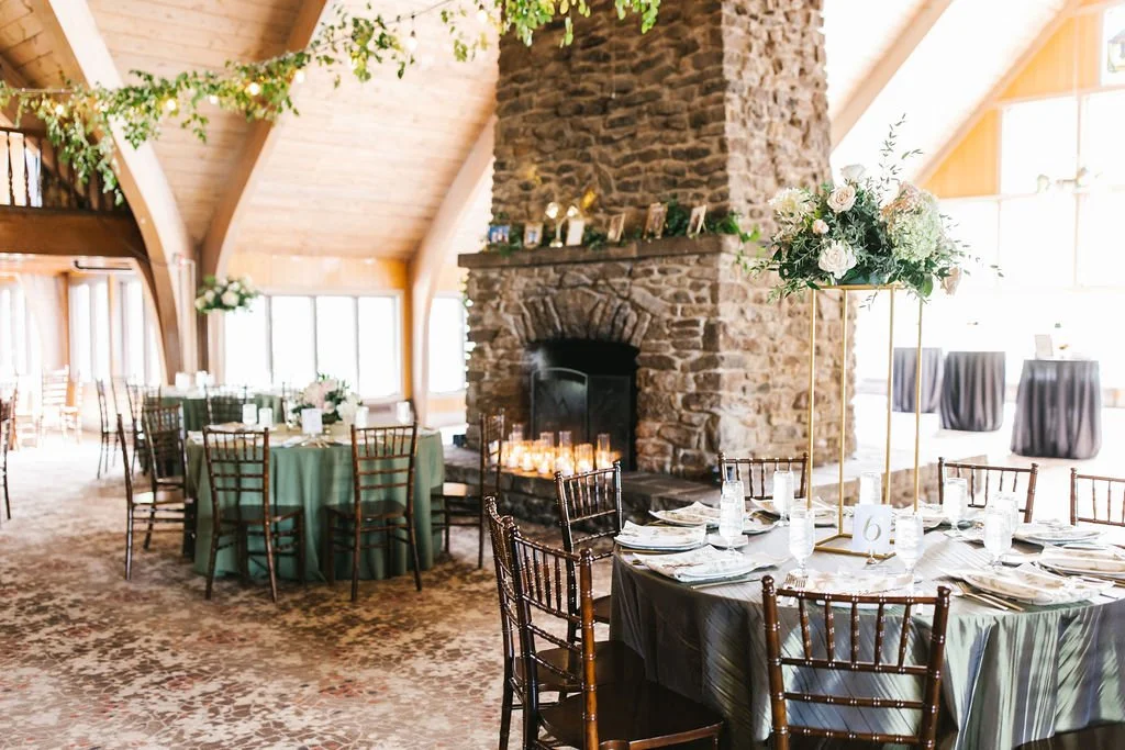 Indoor event space with a large stone fireplace, decorated tables with green and black tablecloths, floral centerpieces, and a high ceiling with wooden beams and string lights.