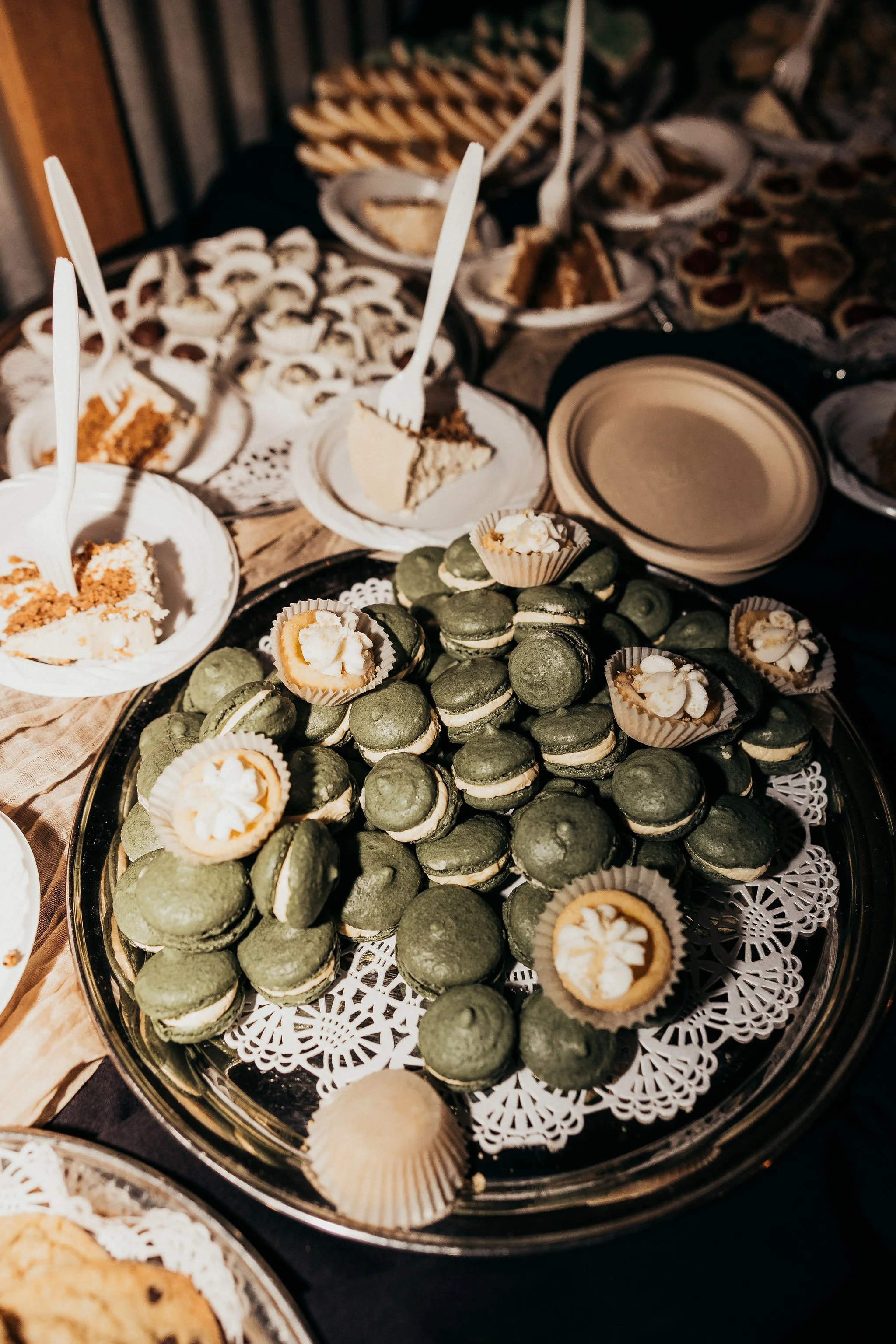 Tray of green macarons with white filling, white and orange petit fours with cream topping, assorted cupcakes, and slices of cake with fruit topping on paper plates.