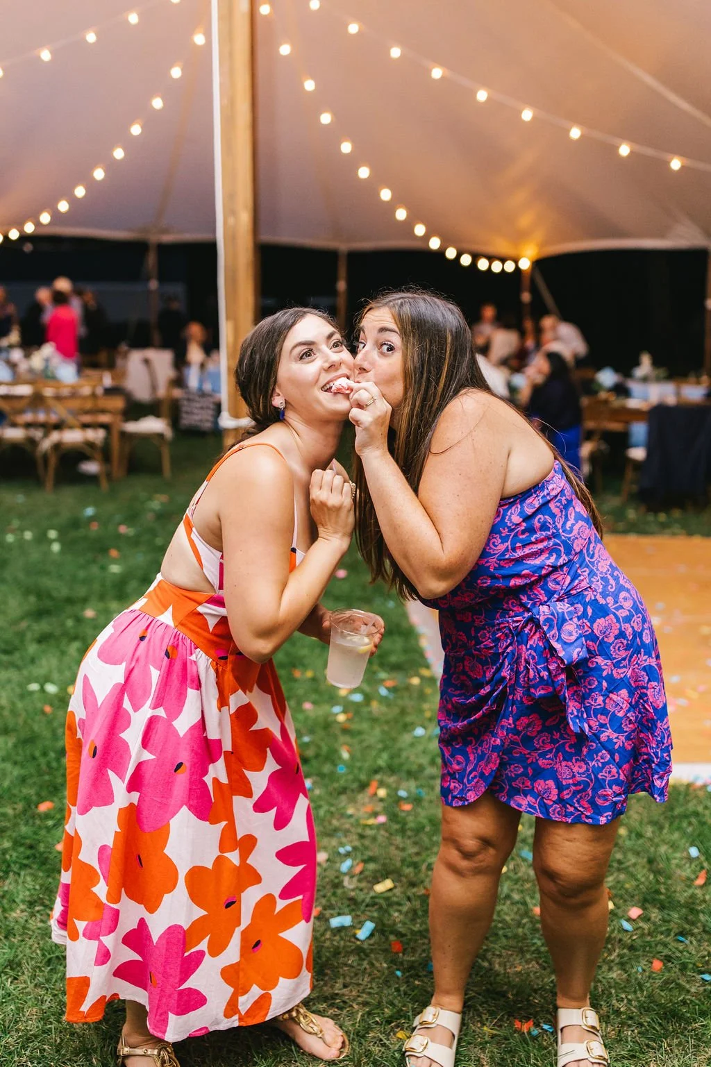 Two women in colorful dresses sharing a playful moment at a festive outdoor event under string lights, with tables and guests in the background.