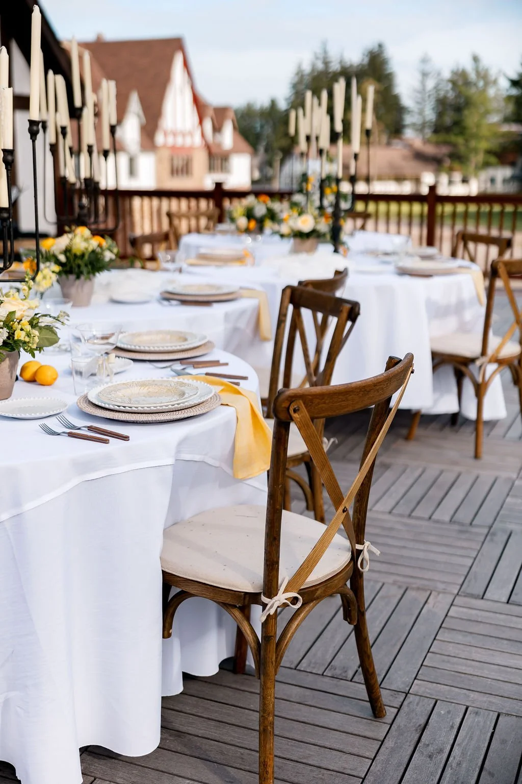 Outdoor dining table set for a formal gathering with white tablecloths, floral centerpieces, and candle holders on a wooden deck, with houses and trees in the background.