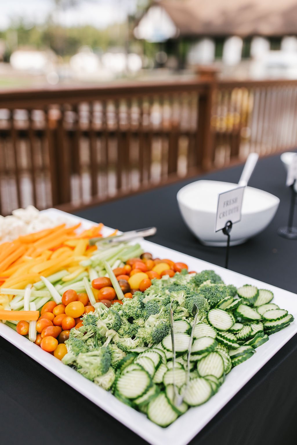 Assorted fresh vegetables including cherry tomatoes, broccoli, sliced cucumbers, celery, shredded carrots, and bell peppers on a white rectangular platter at an outdoor buffet table.