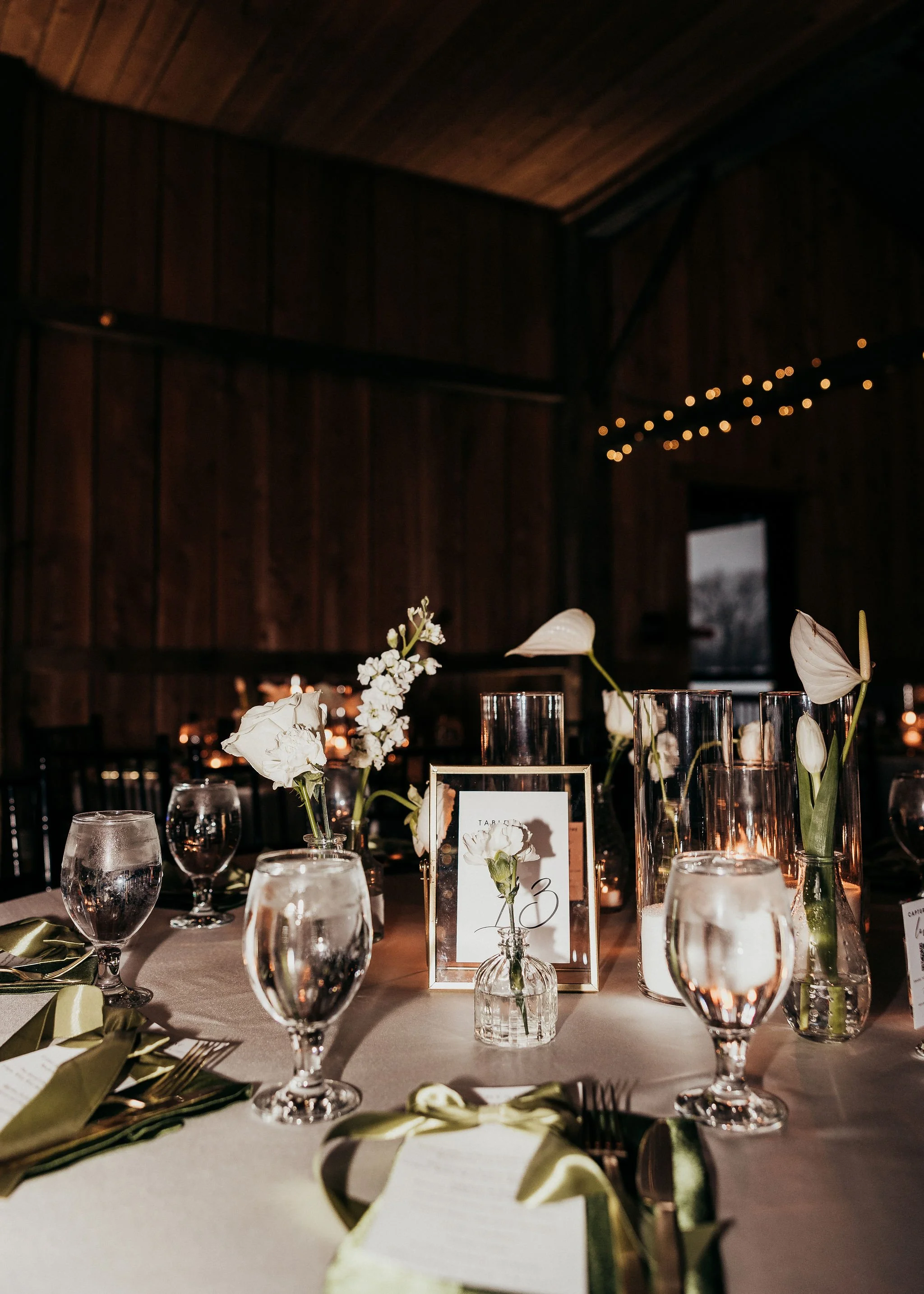 Elegant dinner table setup with flowers and glassware in a dimly lit room with wood-paneled ceiling and string lights