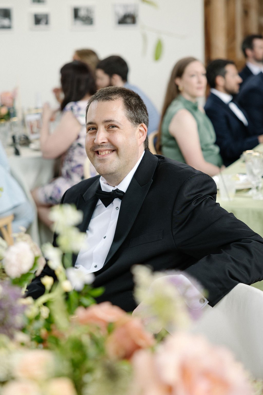 A man in a black tuxedo and bow tie smiling at a formal event, with other guests seated at tables in the background.