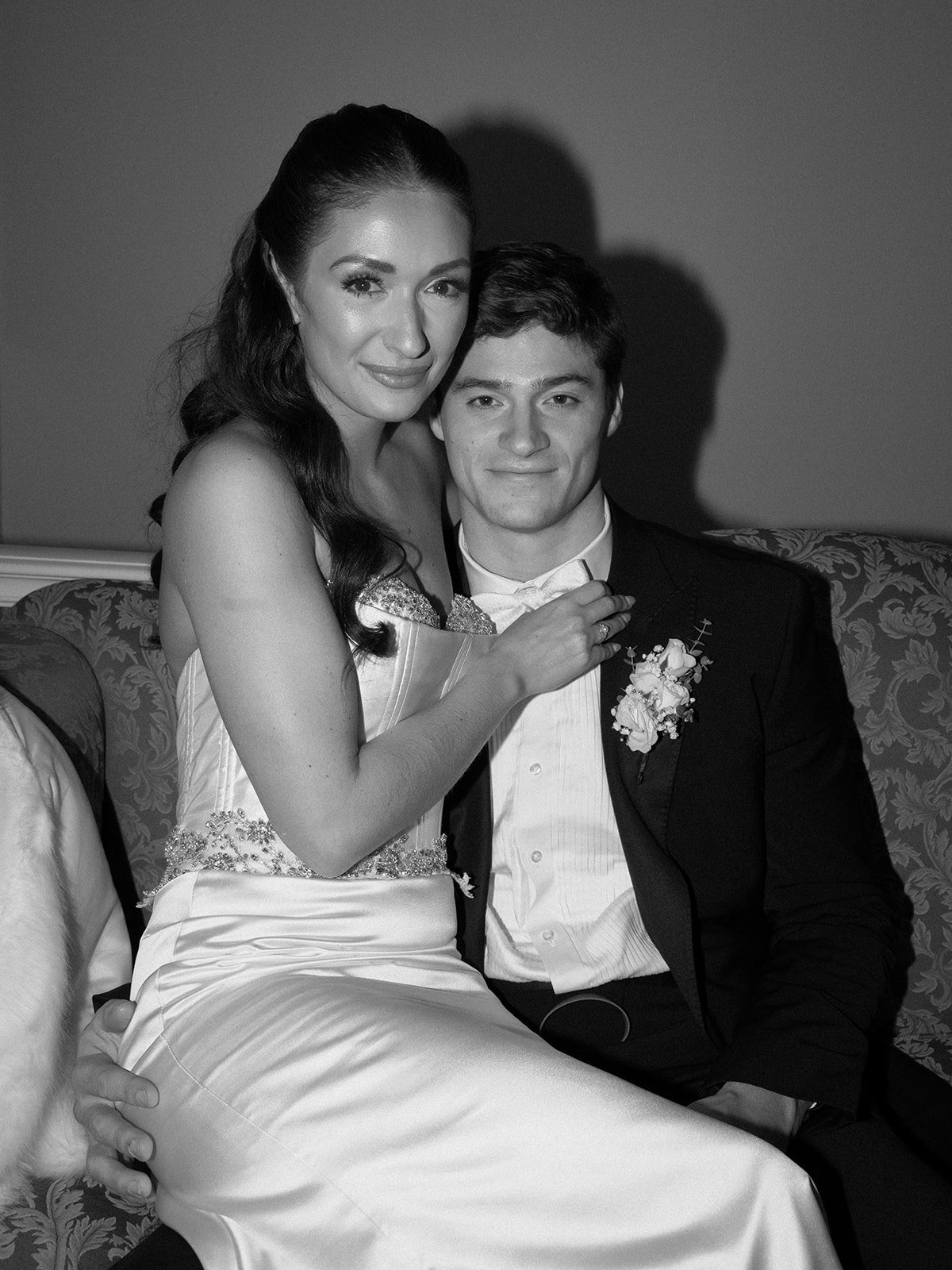 A black and white photo of a newlywed couple sitting close together on a sofa. The bride has long wavy hair and is wearing a strapless wedding gown, while the groom is in a tuxedo with a boutonniere. The bride is smiling softly, and the groom has a s