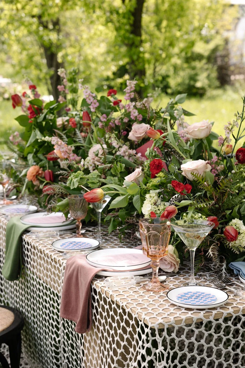 An outdoor dining table decorated with a variety of flowers and pastel-colored glassware, set against a backdrop of green trees and foliage.