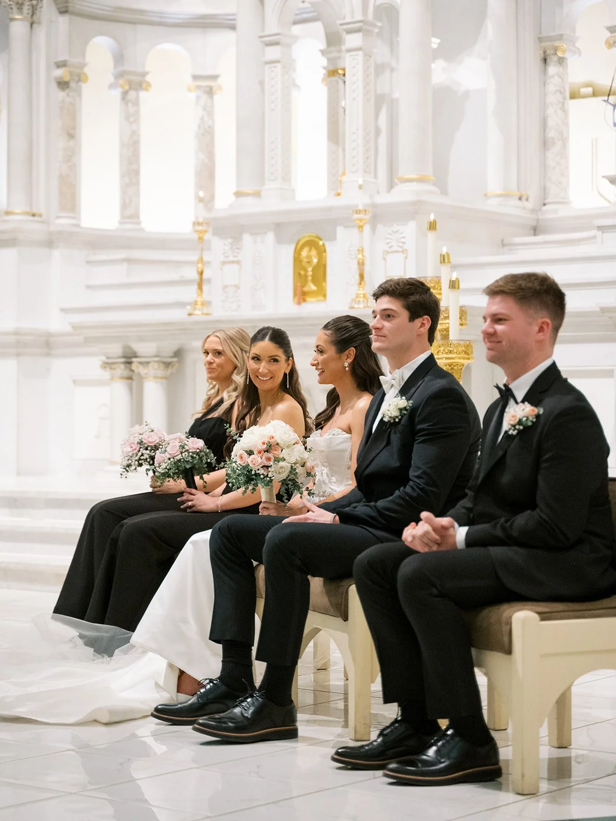 A wedding ceremony with five people sitting on a bench inside a church with white and gold decor, holding floral bouquets, and dressed in formal attire.