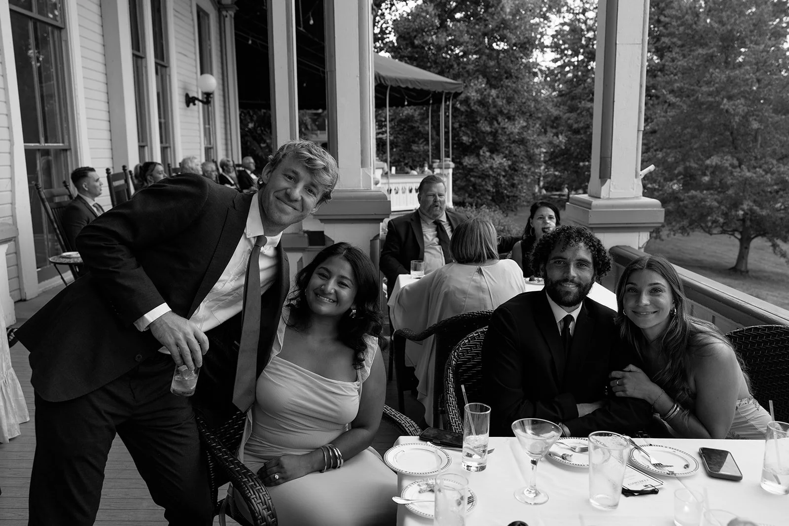 Group of people at a social gathering enjoying drinks on an outdoor porch, with some seated at a table and others standing, in a black and white photo.