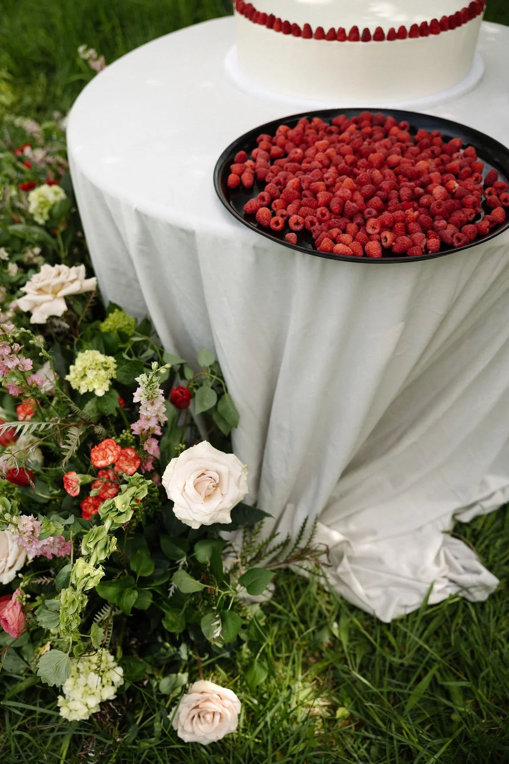 A black plate of raspberries on a white tablecloth, with a decorative cake and colorful flowers surrounding the table outdoors.