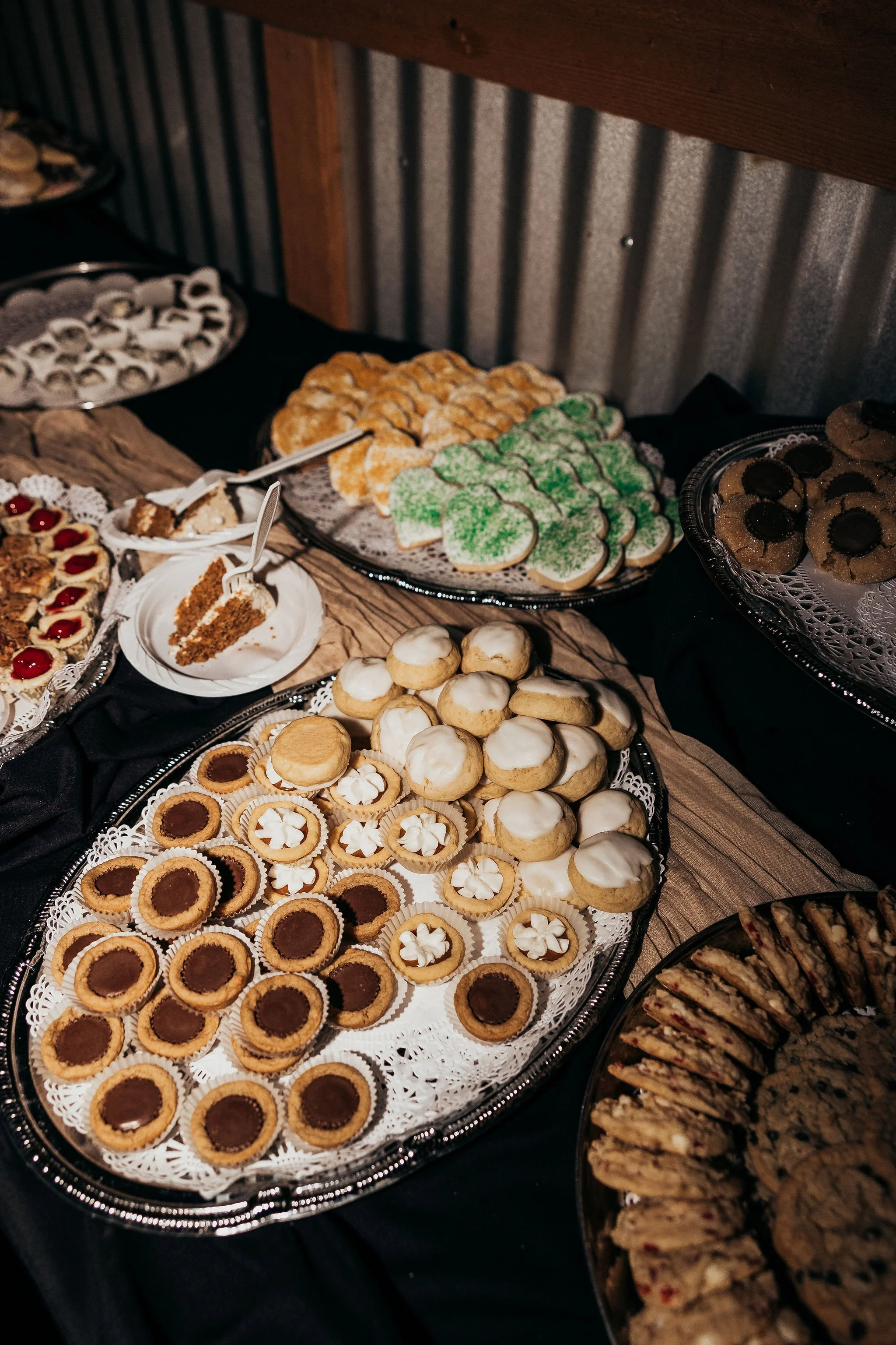 A table with various desserts including cookies, tarts, and pastries displayed on silver trays with doilies, set against a rustic wooden and metal background.