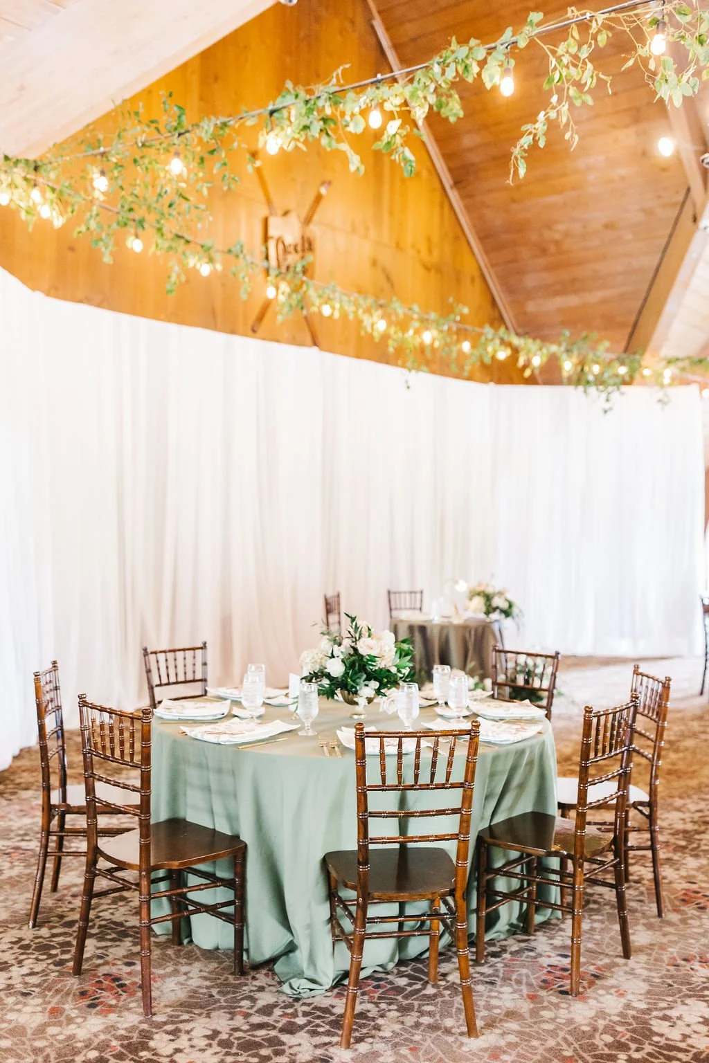 Round banquet table with a green tablecloth, set with plates, glasses, and a floral centerpiece, surrounded by wooden chairs with a wood-paneled ceiling and string lights with greenery above.