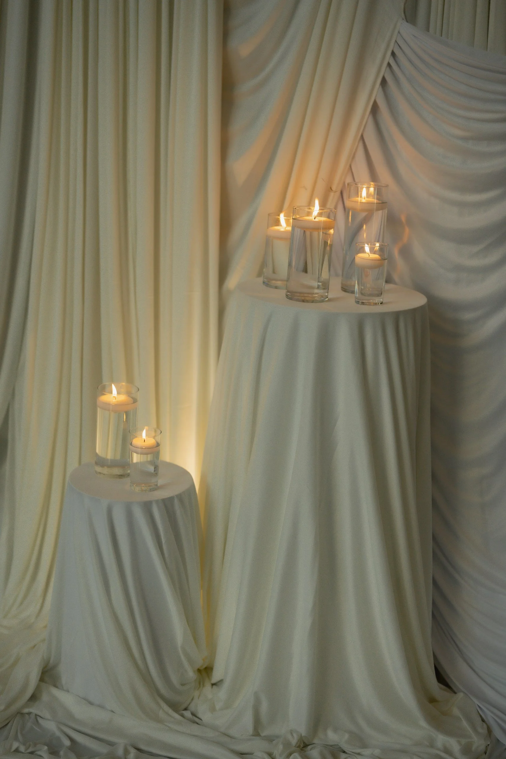 Several candles in glass holders placed on and around two small round tables draped with white cloths, background of cream-colored draped fabric.
