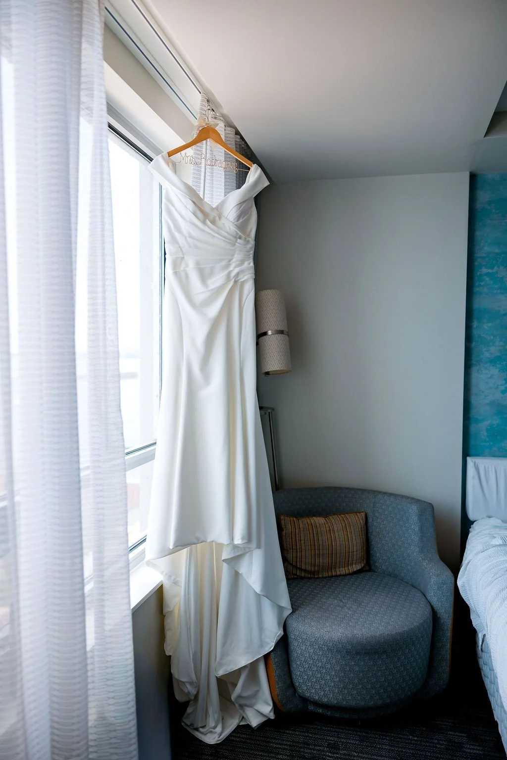 A white wedding dress hanging on a wooden hanger near a window with sheer white curtains in a hotel room.