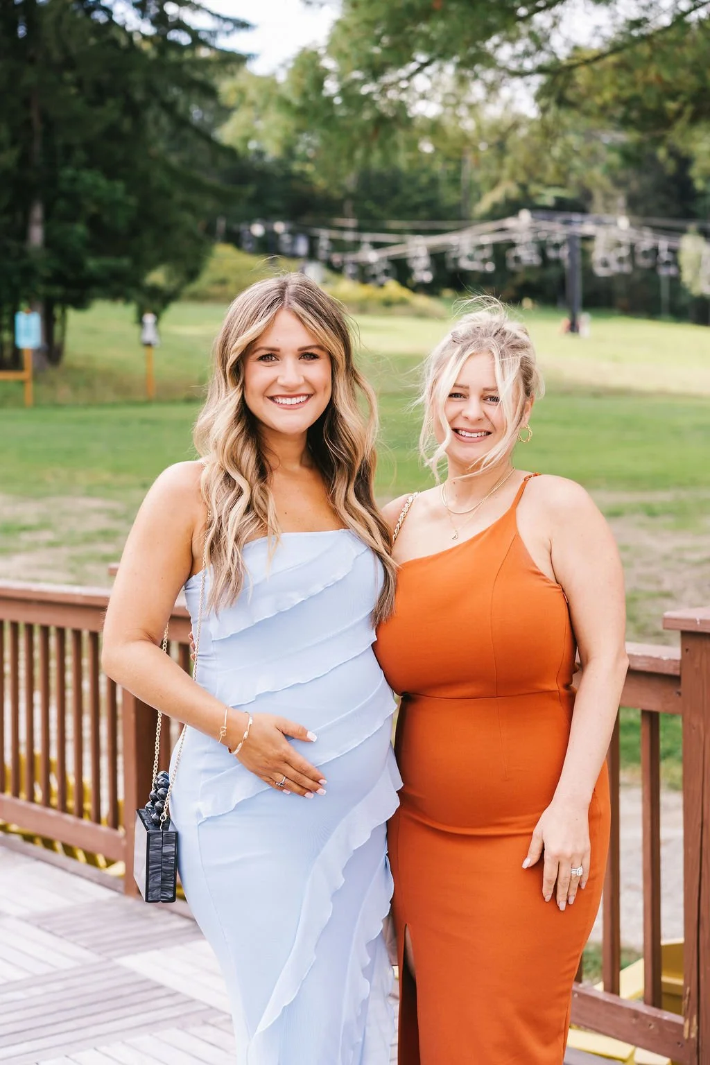 Two women smiling and standing close together outdoors on a wooden deck, with trees and green fields in the background. One woman is wearing a white dress, and the other is wearing an orange dress.