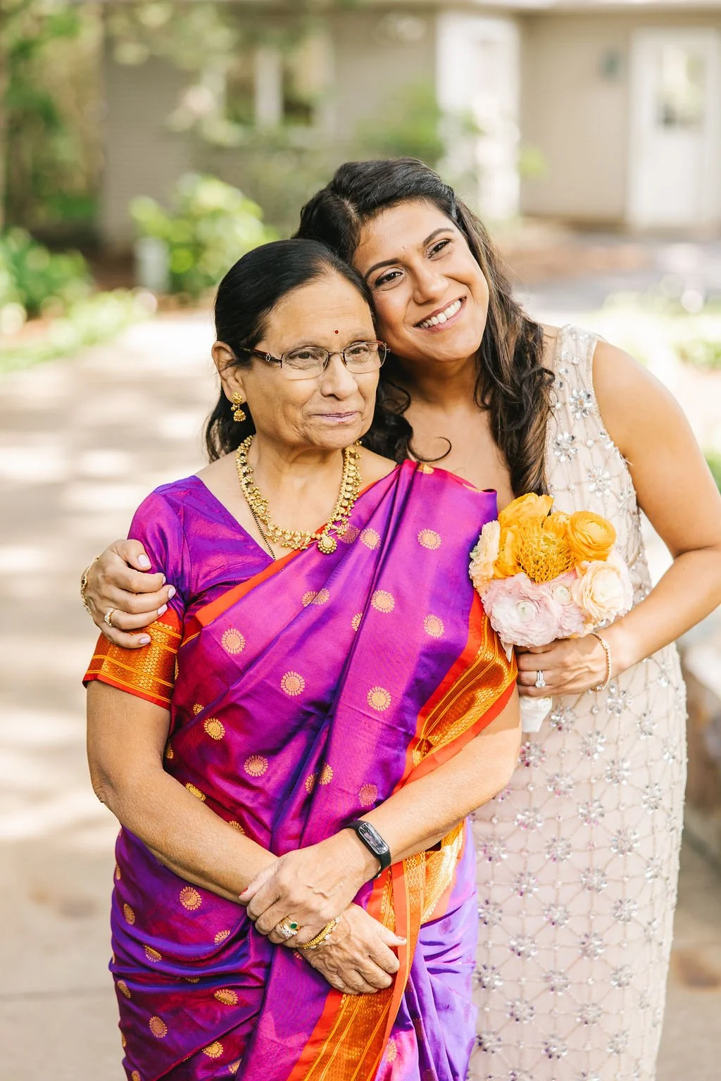 A young woman and an elderly woman are standing outside, smiling and posing for a photo. The young woman is holding a bouquet of flowers, and both women are dressed in traditional Indian attire. The older woman is wearing a purple and orange saree, a