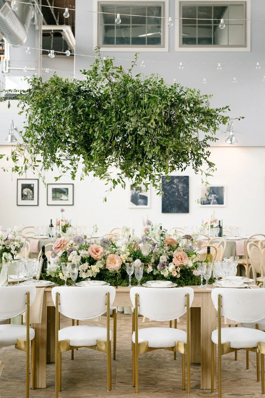 Elegant dining setup with a large floral centerpiece, white chairs with gold accents, and a lush green hanging plant overhead in a bright, modern event space.