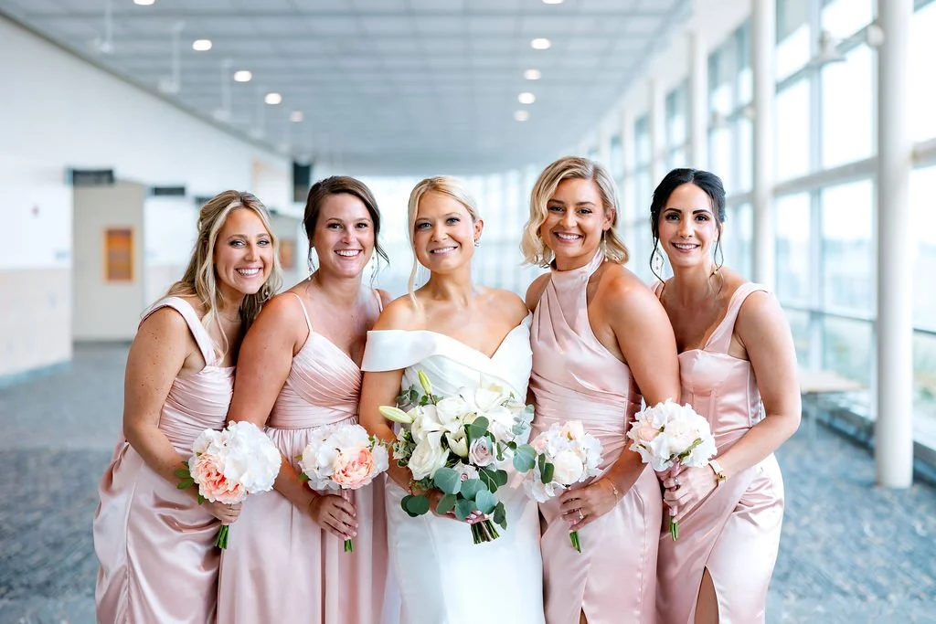 A bride with blonde hair in a white wedding dress holding a bouquet standing with five bridesmaids in light pink dresses, all holding bouquets, inside a modern, glass-walled building.