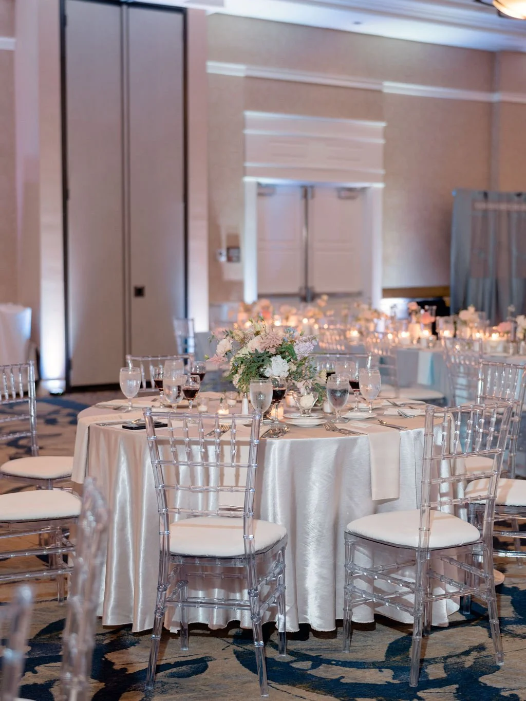 Elegant banquet table set with clear chairs, floral centerpiece, water glasses, and candles in a decorated event hall.