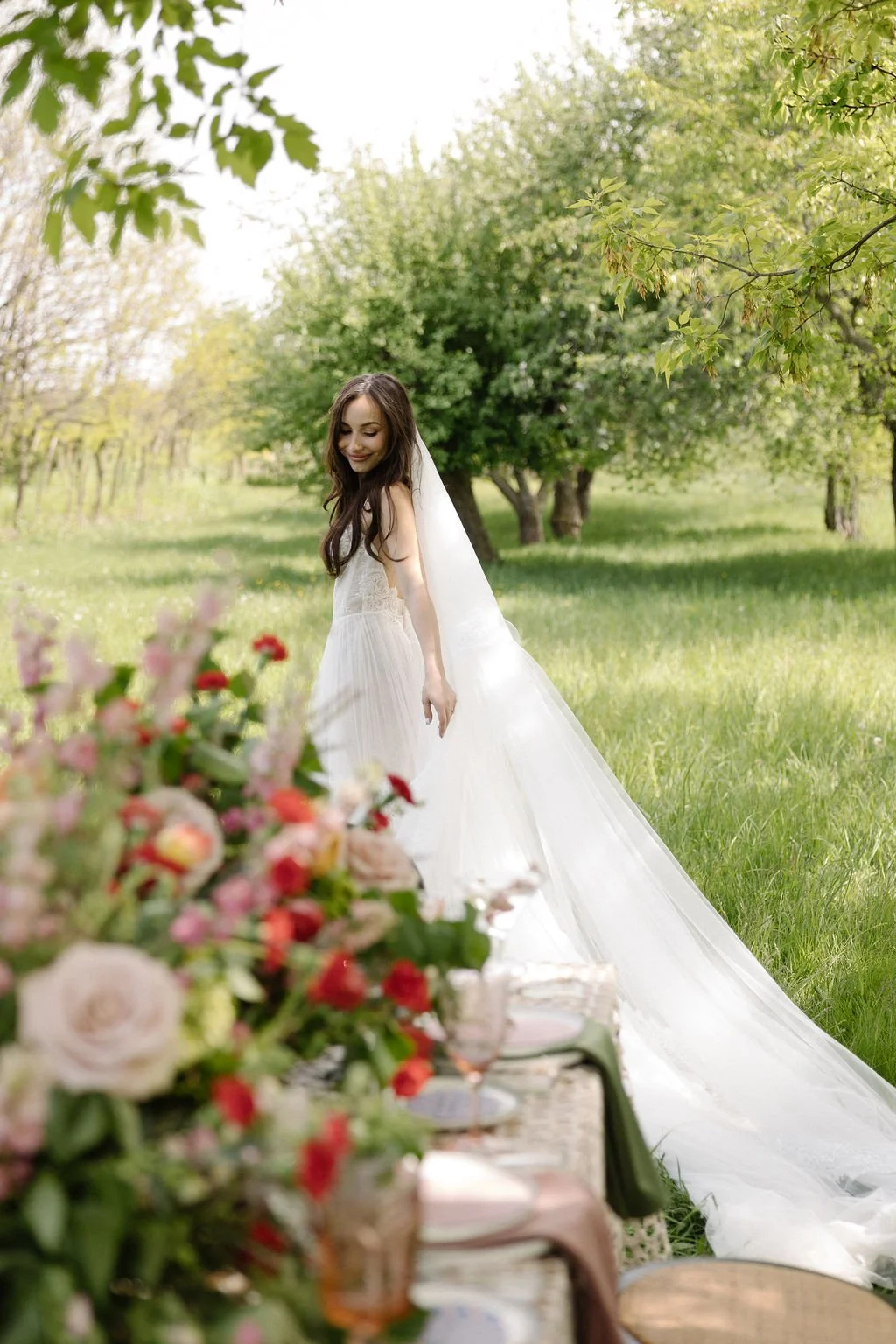 A bride in a long white wedding dress with a veil, smiling and looking down in a lush green outdoor setting with trees and a decorated table with flowers in the foreground.