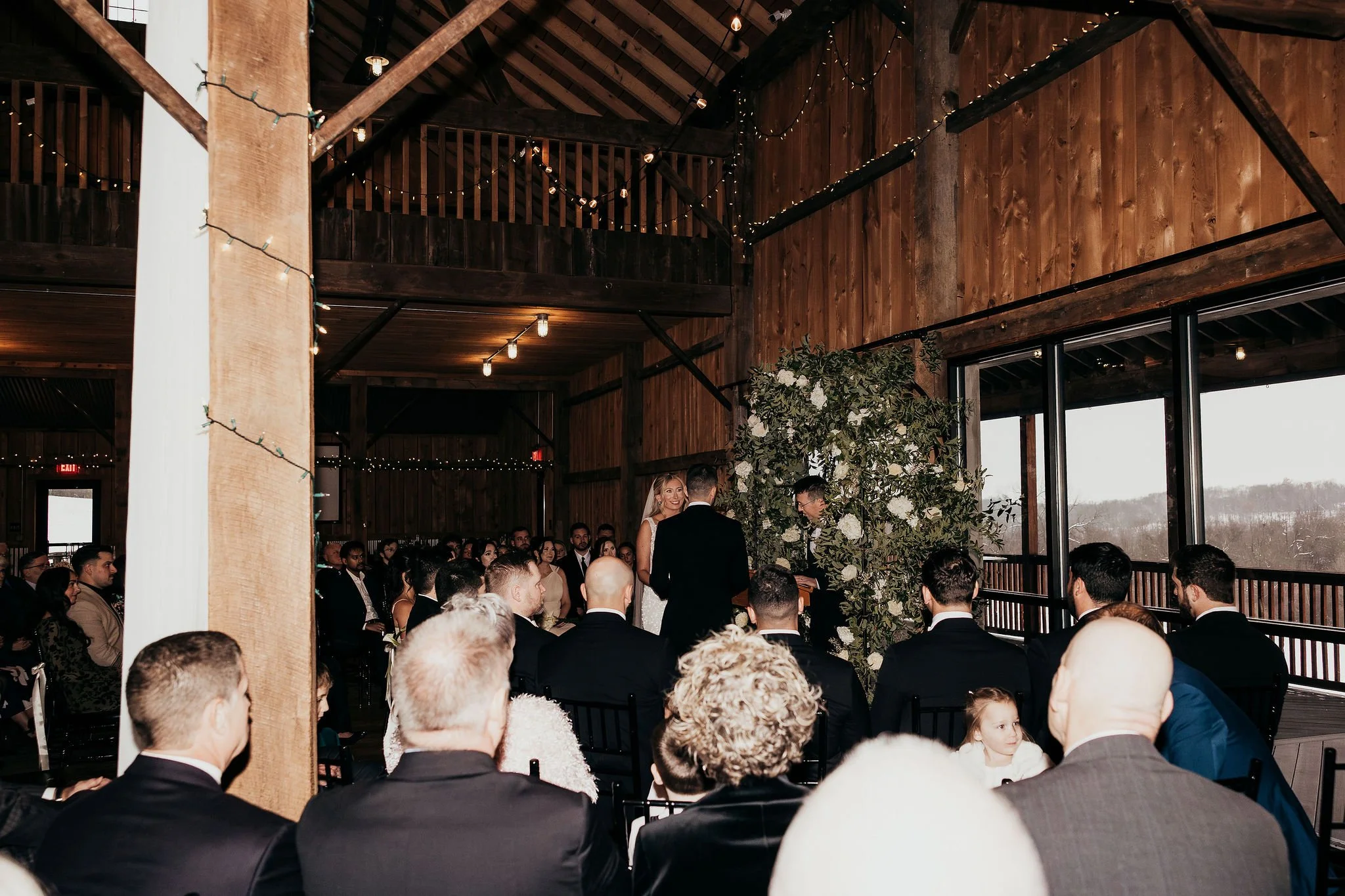 A wedding ceremony taking place inside a rustic wooden venue with string lights, guests seated, and a couple exchanging vows.