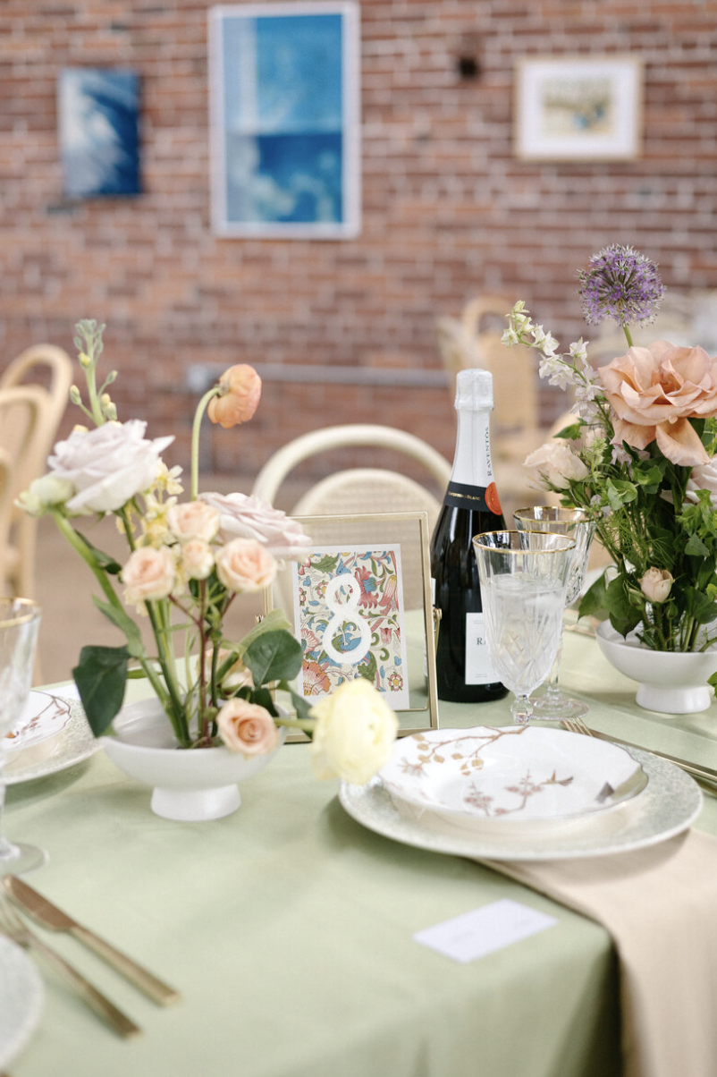 Elegant table setting with floral arrangements, champagne bottle, and glassware at an outdoor event with a brick wall background.