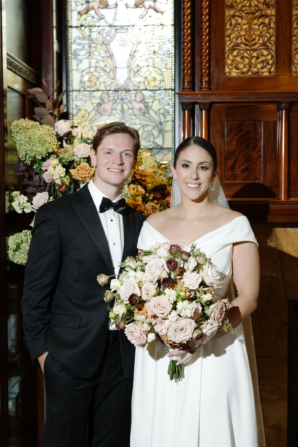 A bride and groom smiling at the camera during their wedding, standing in front of a stained glass window and a floral arrangement.