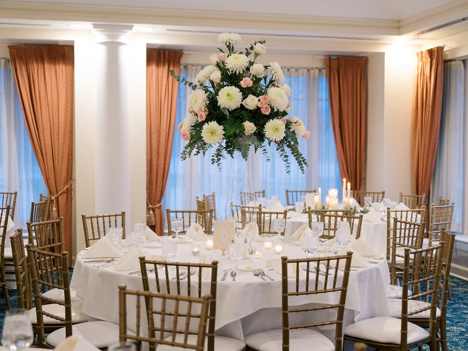 Elegant banquet hall with round tables covered in white tablecloths, gold chairs, and a large floral centerpiece with white and pink flowers and greenery.