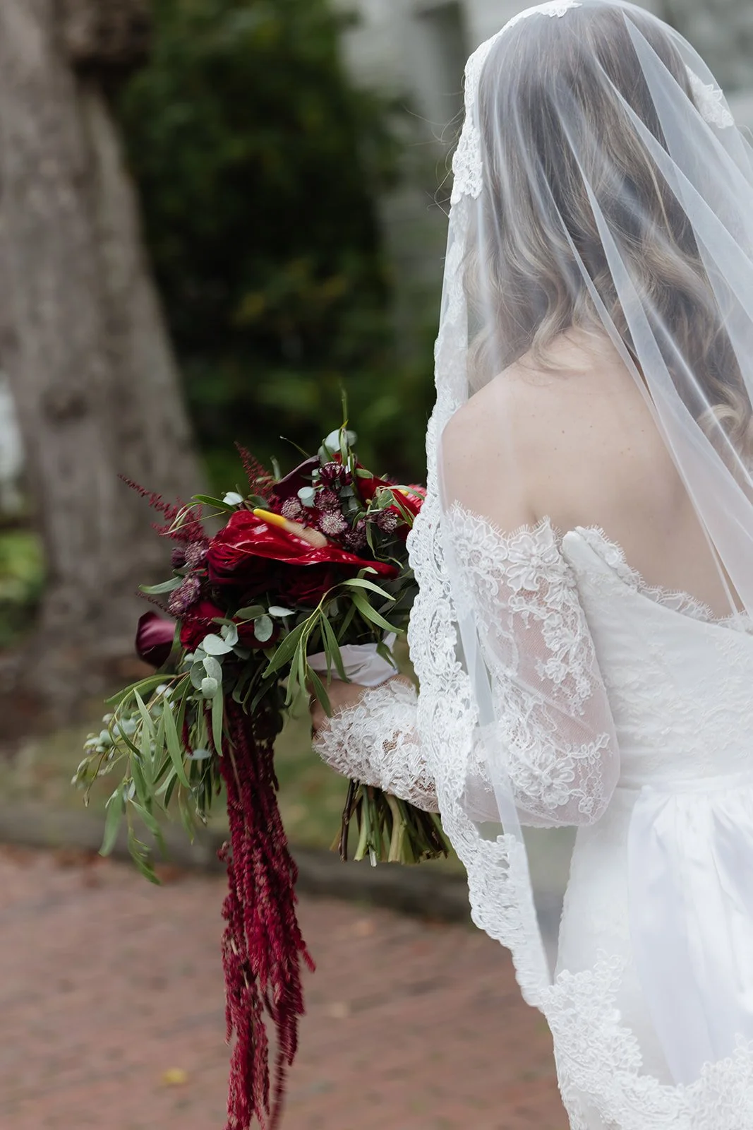A bride in a white lace wedding dress with a veil holding a bouquet of dark red and burgundy flowers with greenery, outdoors near a tree.