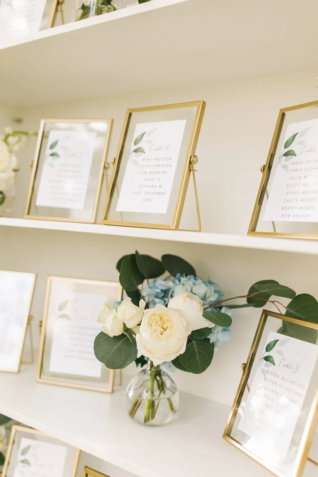 Gold picture frames with seating arrangements and a clear vase with white and blue flowers on a white shelf.