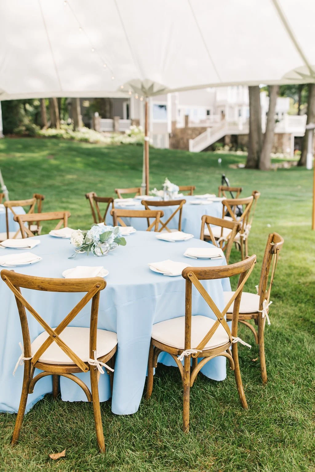 Outdoor event setup with wooden chairs and round tables covered with light blue tablecloths under a tent, with a grassy yard and a house in the background.