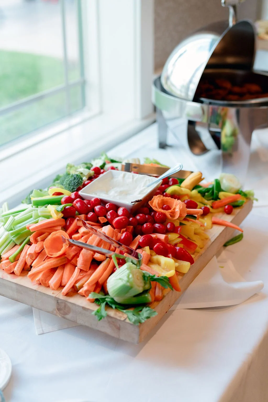 Colorful vegetable platter with cherry tomatoes, celery, carrots, broccoli, yellow bell peppers, and cucumbers, with a bowl of ranch dip on top. In the background, a slow cooker is visible on a kitchen counter near a window.