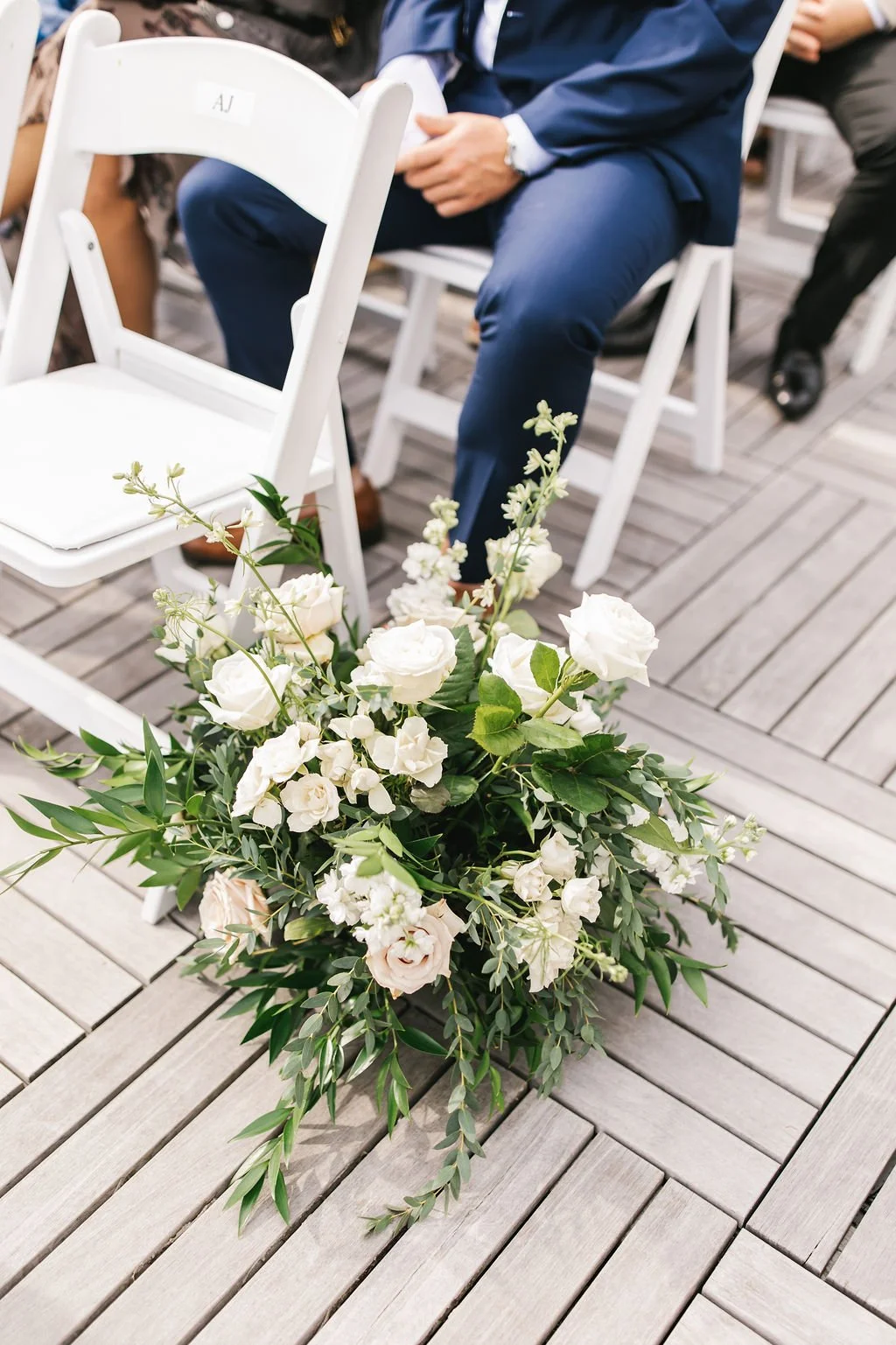 White floral arrangement on a wooden floor at a wedding or event, with people seated behind it.