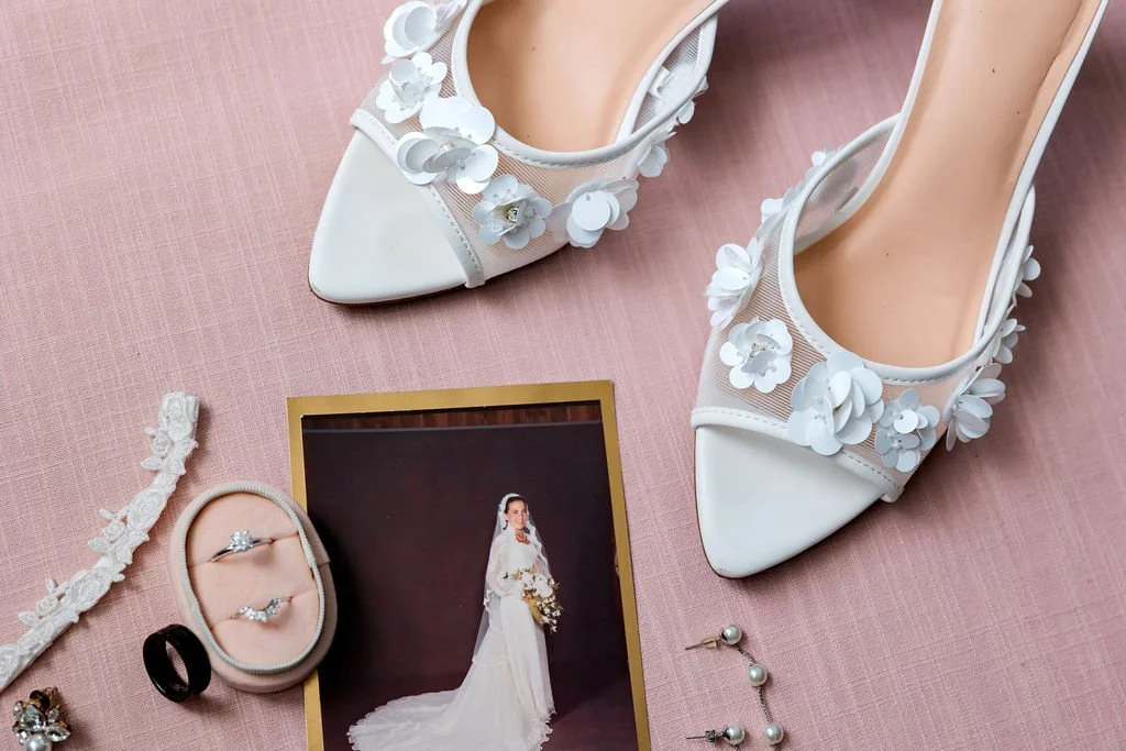 White bridal shoes with floral decorations, a wedding dress photo, rings in a box, earrings, and a bracelet on a pink surface.