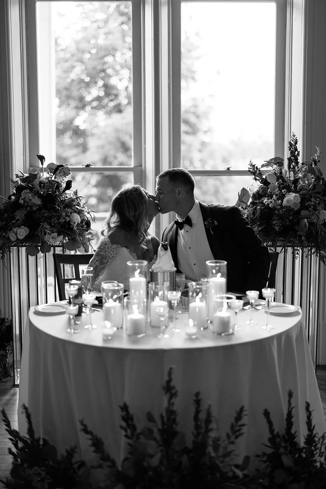 A black and white photo of a bride and groom sharing a kiss at their wedding reception, sitting at a table decorated with candles and flowers, with large windows and natural light in the background.
