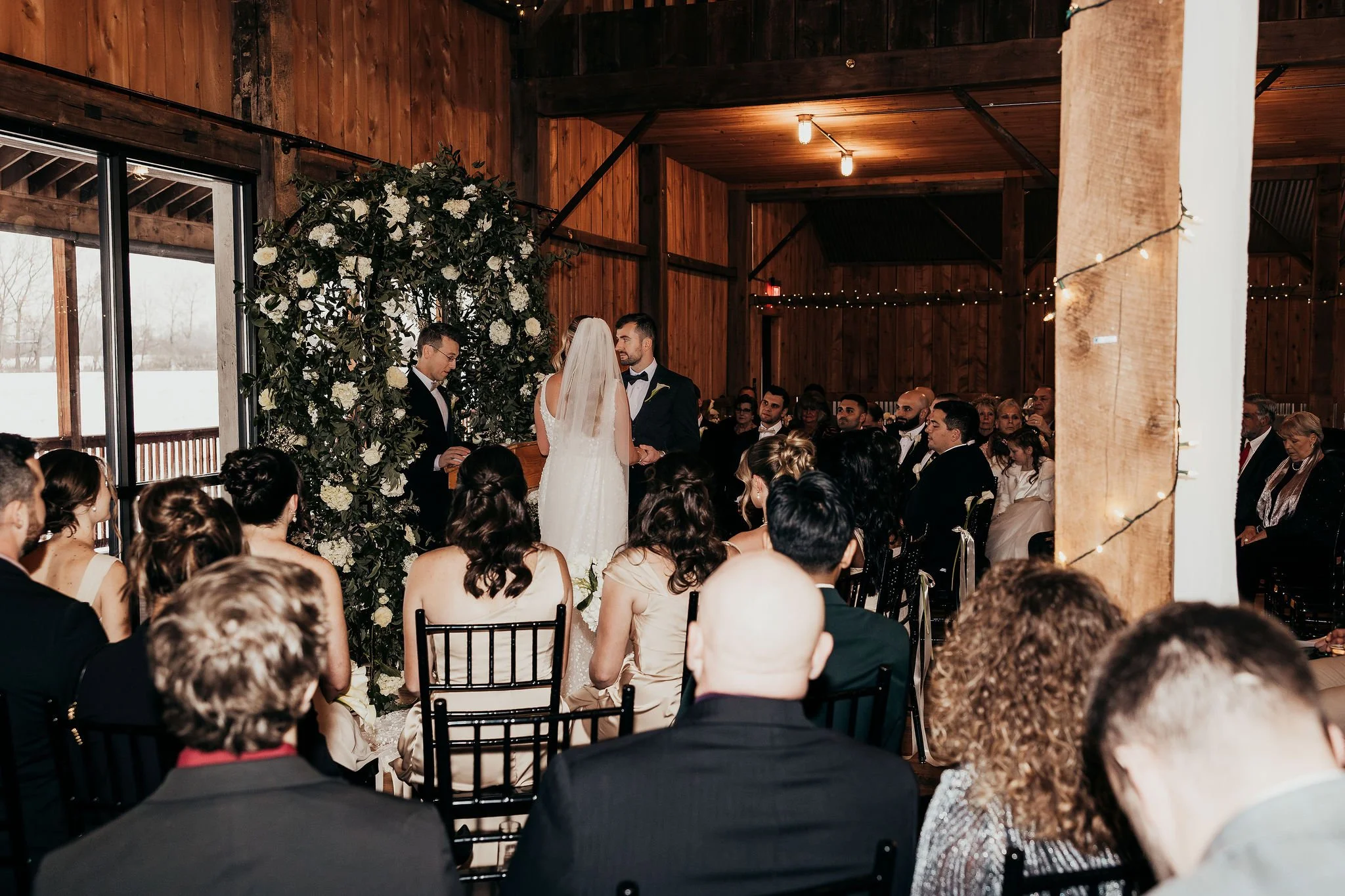 A wedding ceremony taking place inside a wooden venue with a bride and groom standing before an officiant. The bride is wearing a white dress and veil, while the groom is in a black tuxedo. Guests are seated and watching the ceremony, and a floral ar