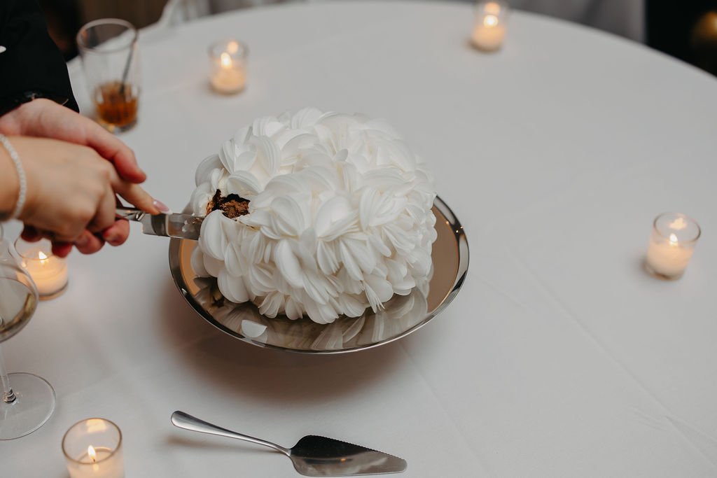 A person cutting a piece from a white frosted cake with chocolate filling on a silver platter, surrounded by lit candles on a round table covered with a white tablecloth.