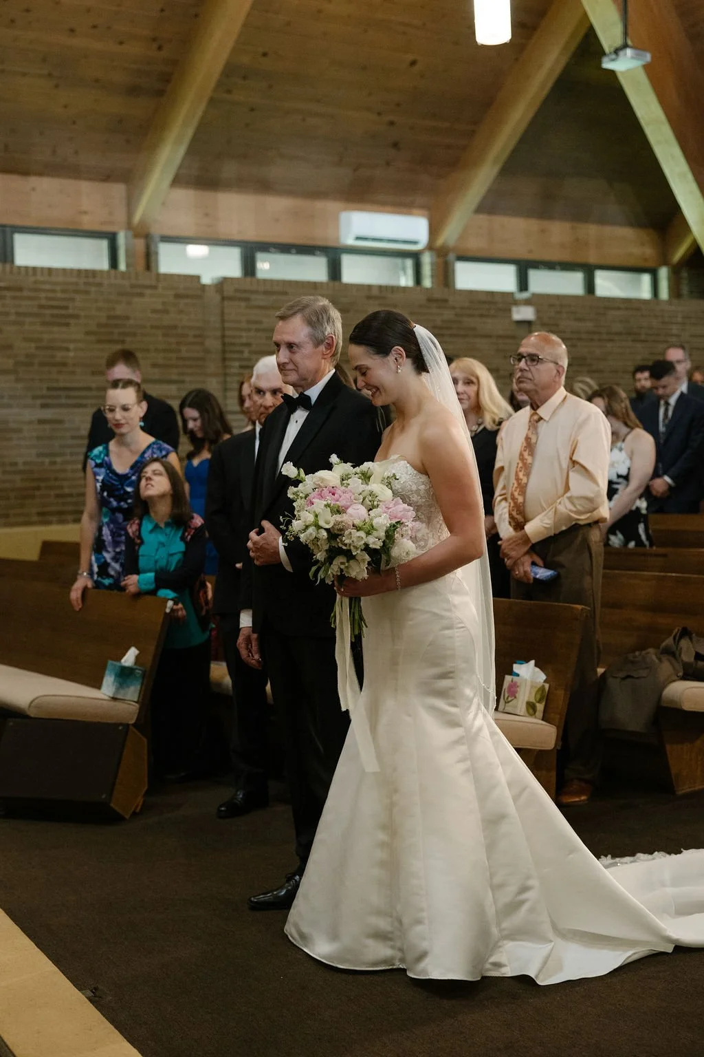 A bride in a white wedding gown holding a bouquet of pink and white flowers, walking down the aisle with an older man in a black tuxedo. Guests stand and sit in pews, watching in a wooden church with a vaulted ceiling.