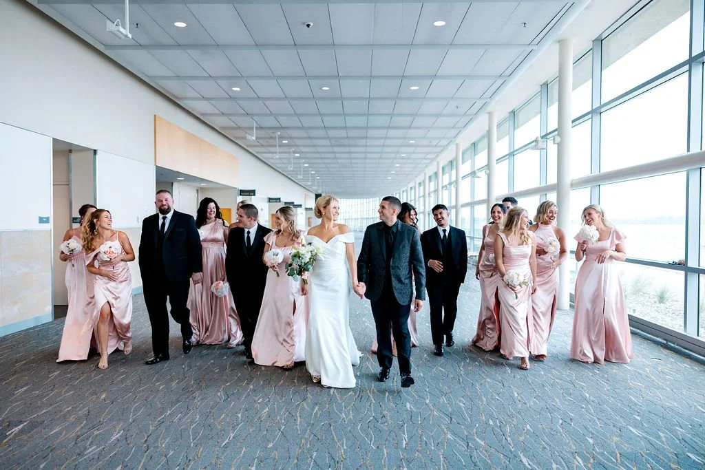 Bride and groom with wedding party walking through a spacious, modern corridor with large windows and natural light.