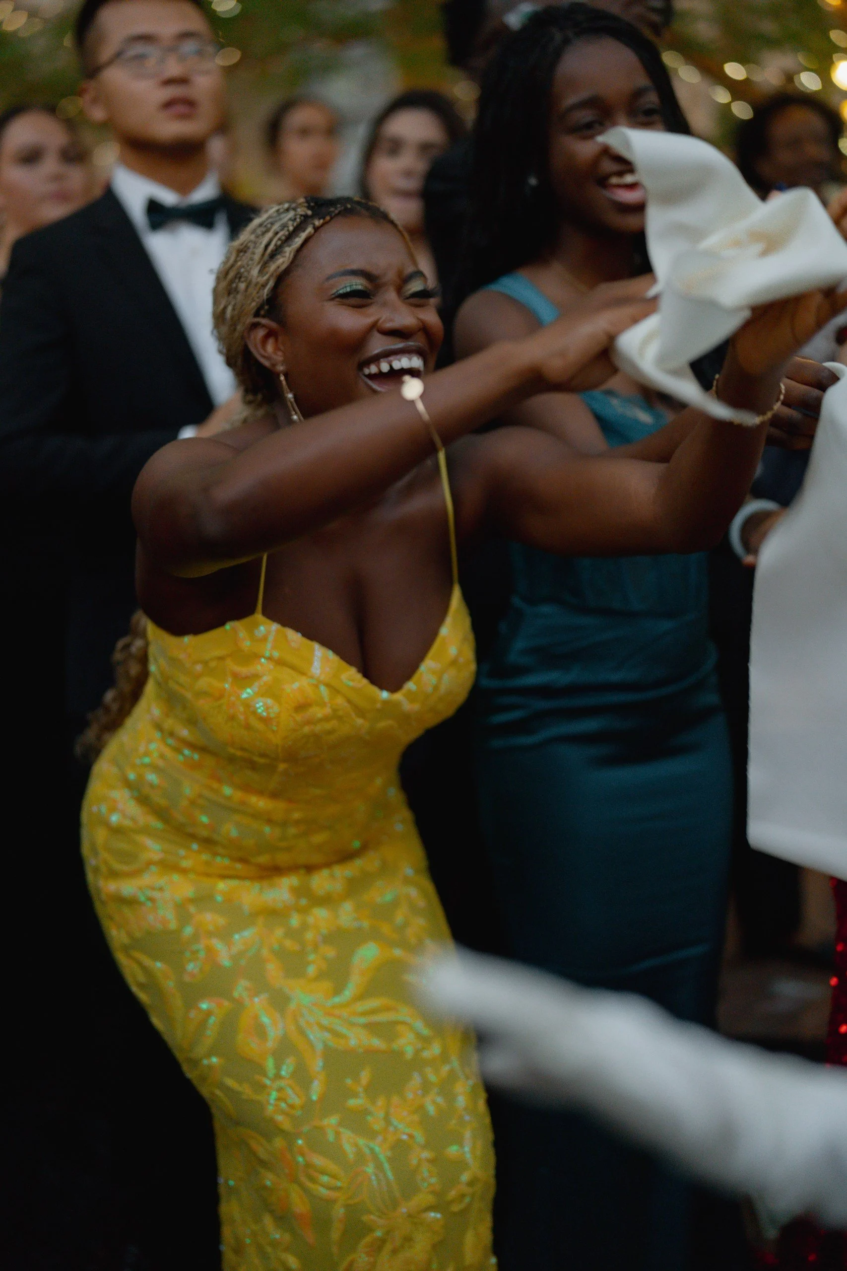 People at a celebration or party, holding a white cloth or napkin, smiling and dancing, with some dressed formal and others in colorful dresses.