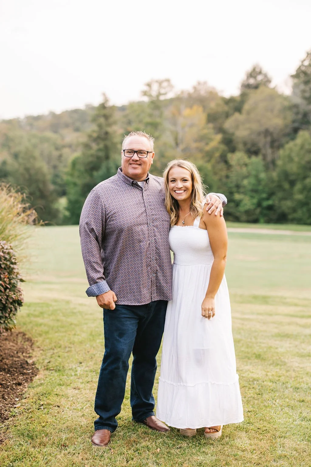 A man and a woman standing outdoors on a grassy area with trees in the background. The man is wearing glasses, a patterned button-down shirt, and dark jeans. The woman is dressed in a white sleeveless dress, and they are smiling with the man's arm ar
