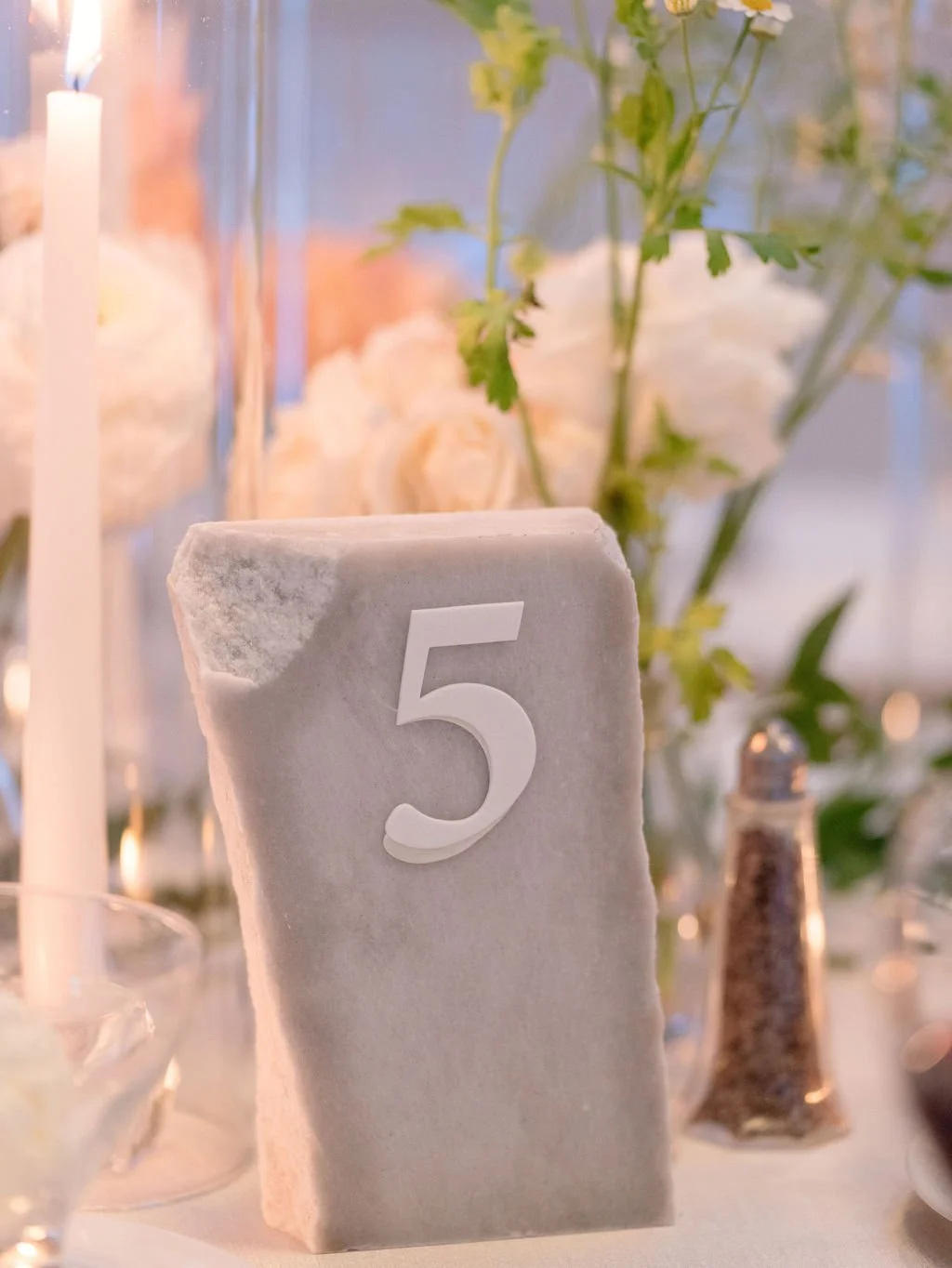 Table centerpiece with a white marble block displaying the number 5, surrounded by flowers, a tall candle, a salt shaker, and other table items, at an elegant event.