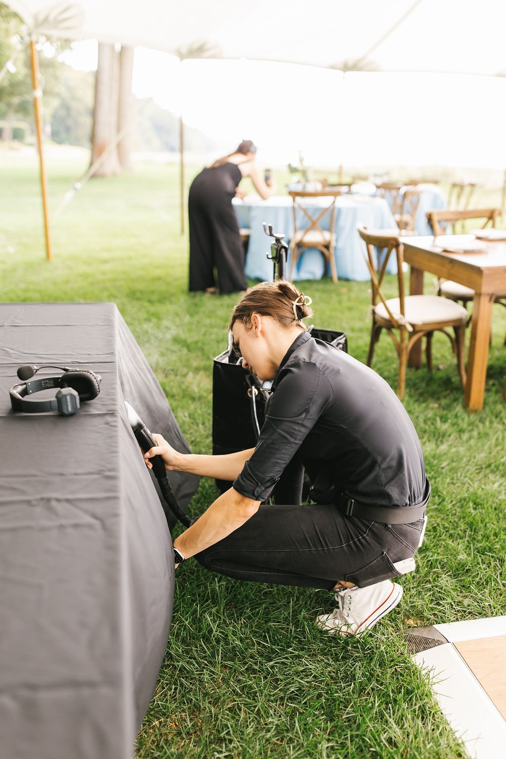 Woman cleaning a table at an outdoor event under a tent, with chairs and a woman taking photos in the background.