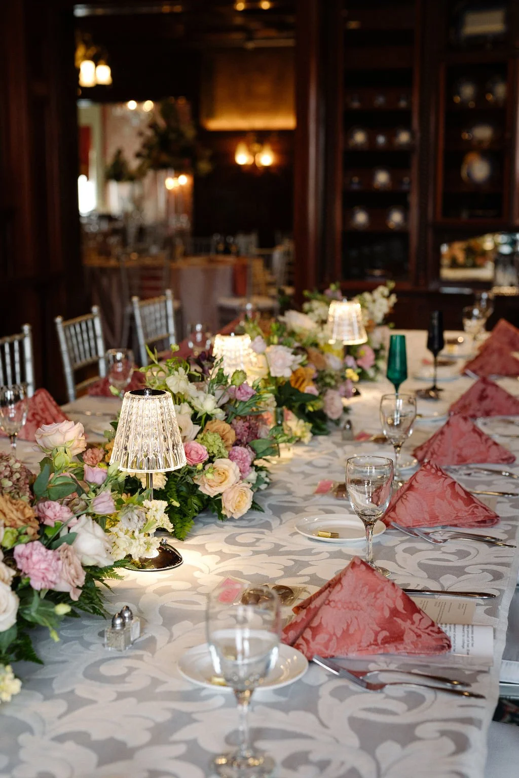 Elegant dining table with floral centerpiece, pink napkins, glassware, and small lamp lights, in a restaurant or banquet hall.