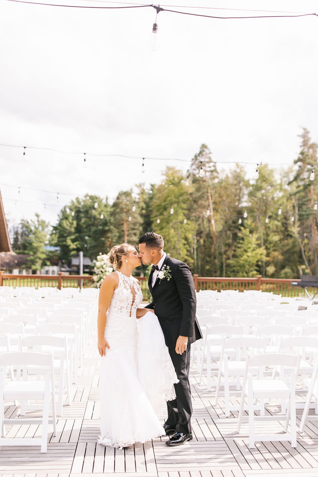 A bride and groom kiss outdoors during their wedding ceremony, with empty chairs arranged behind them and string lights overhead.