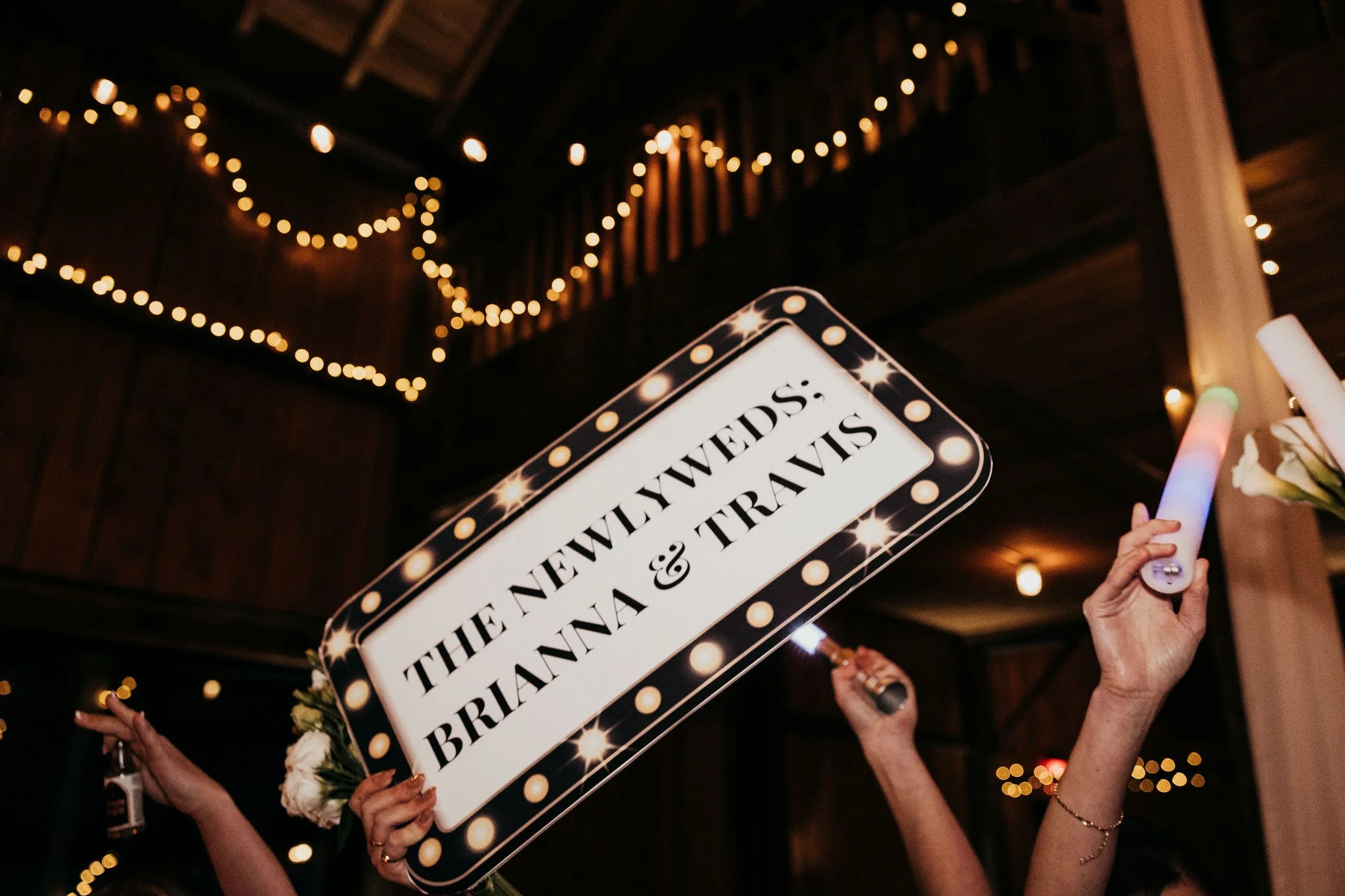 People holding a sign that reads 'The new brides; the anniversary &' with a background of string lights and a wooden ceiling, likely at a celebration or wedding event.