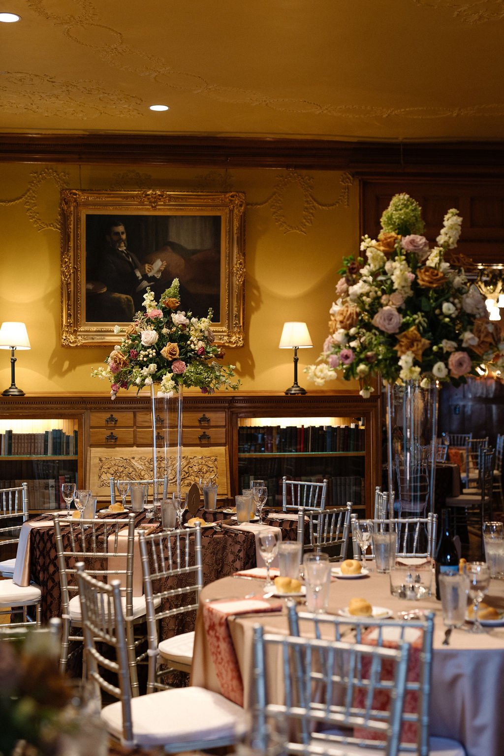 Elegant dining room with round tables, floral centerpieces, silver chairs, and framed portrait on the wall.