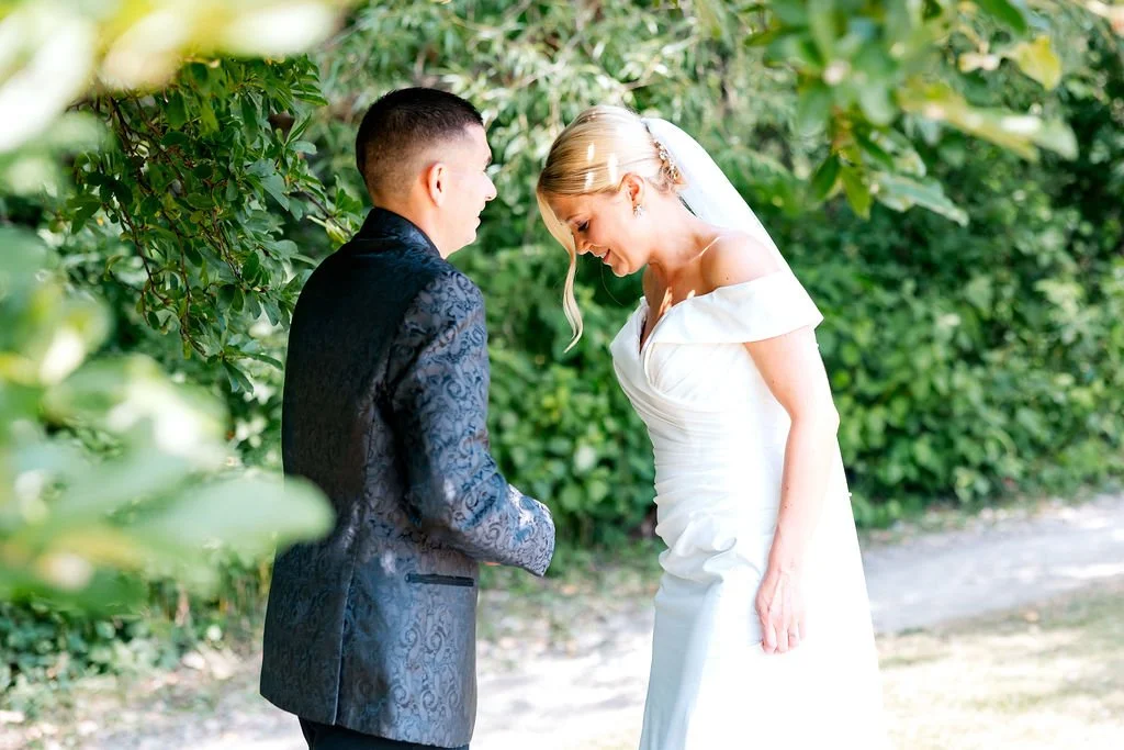 A bride and groom standing outdoors during a wedding ceremony, with the bride smiling and looking down, and the groom facing her, surrounded by greenery.