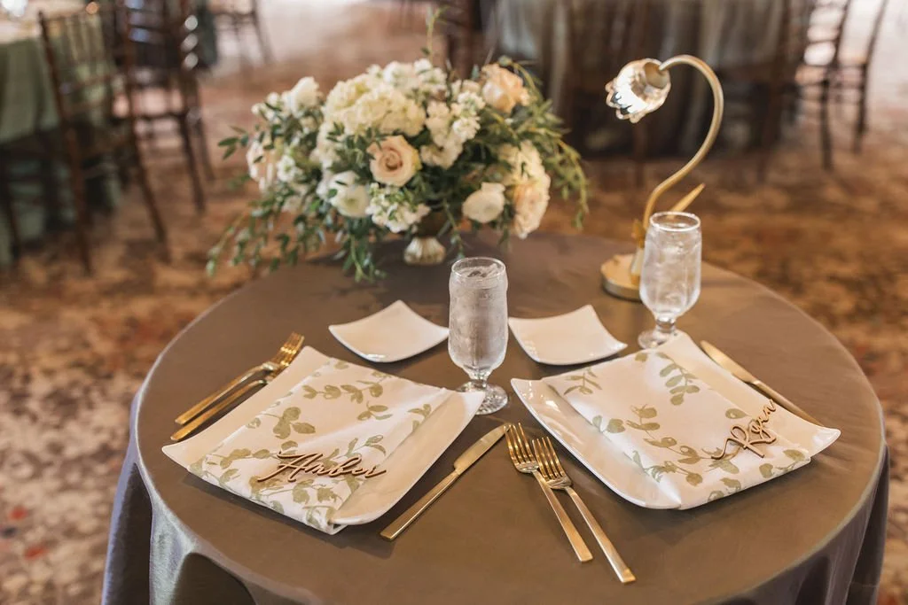 A round dining table set for two with a floral centerpiece, two glasses of water, gold cutlery, and decorative napkins with gold lettered place cards that say 'Aubrey' and 'Reed', under a warm lighting in a venue with chairs and a patterned carpet.
