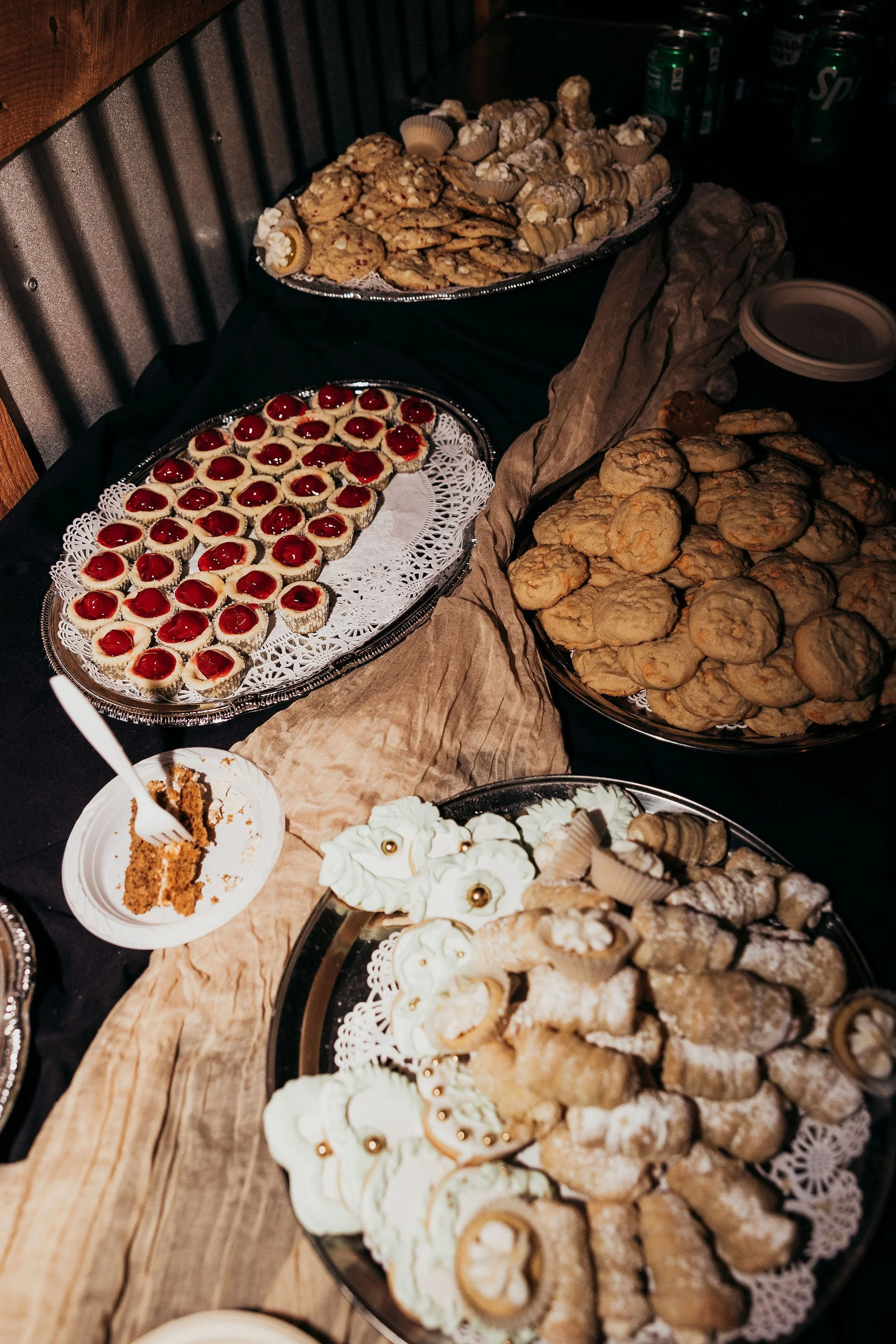 Assorted cookies and desserts on silver trays with doilies, including cherry-topped cookies, nut cookies, and sugar cookies, arranged on a rustic wooden table. Beverages are in the background.