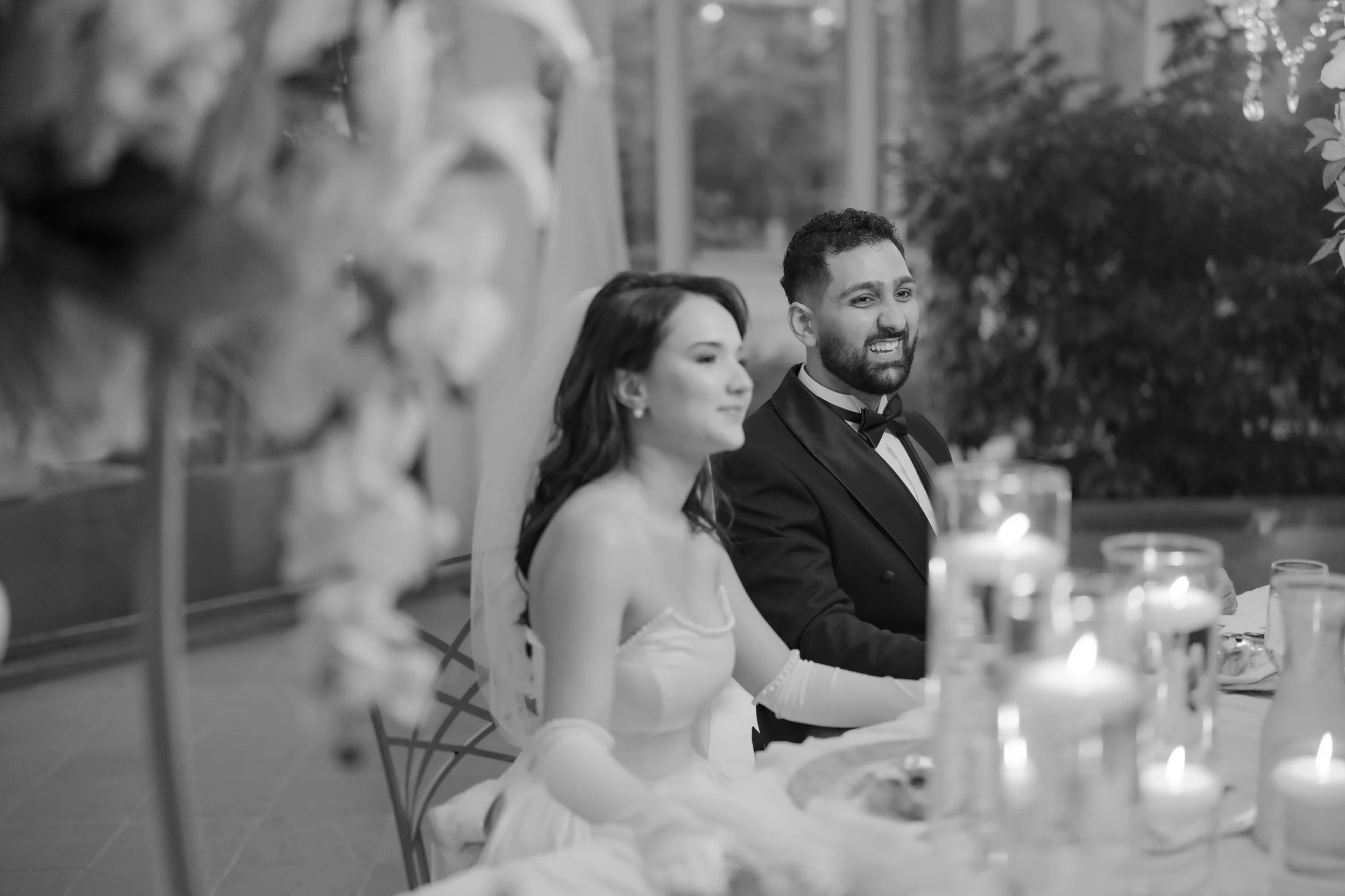 A black and white photo of a man and a woman dressed in formal attire, seated at a table during a celebration, with blurred background and guests.
