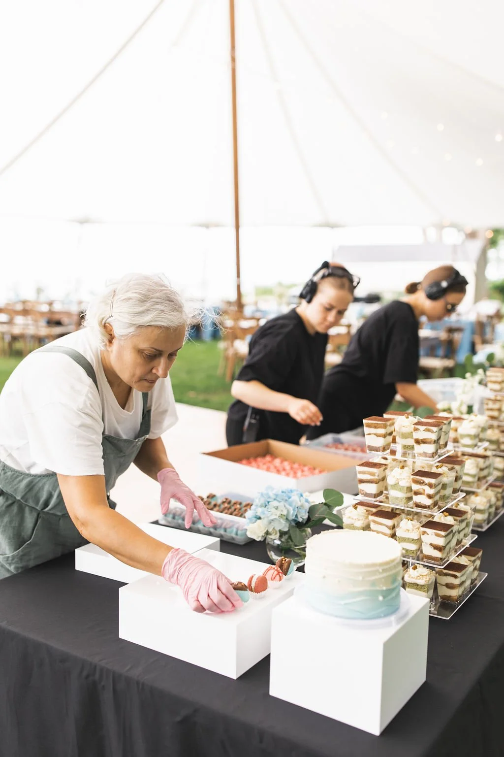 Three women working at a dessert table under a large outdoor tent, arranging and serving desserts including cakes and layered parfaits.